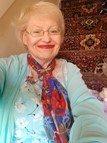 Deb Smith is a VDRP member. She is a white woman with blonde hair. She is smiling, wearing red lipstick and glasses. She has a red patterned scarf around her neck and wears a pale blue floral top, with a pale blue cardigan.