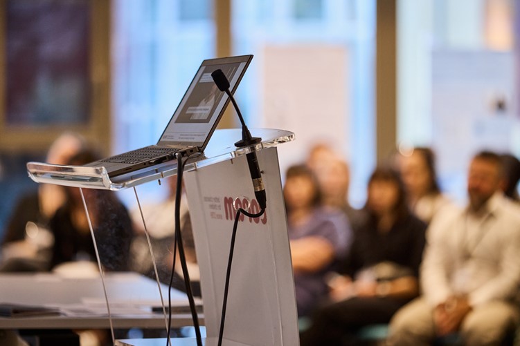 A photo of a lecturn on a stage, with a microphone and a laptop, with an audience waiting for a presentation