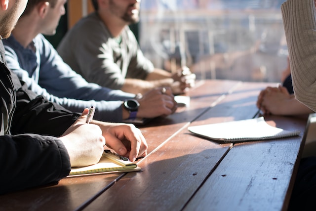 Several people sit around a table, their hands on the table on notebooks and holding pens